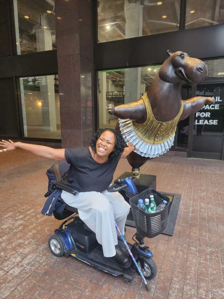 A photo of Lizzie Kiama, a Kenyan disability rights activist, posing in front of a statue of a hippo in a ballerina costume and pose